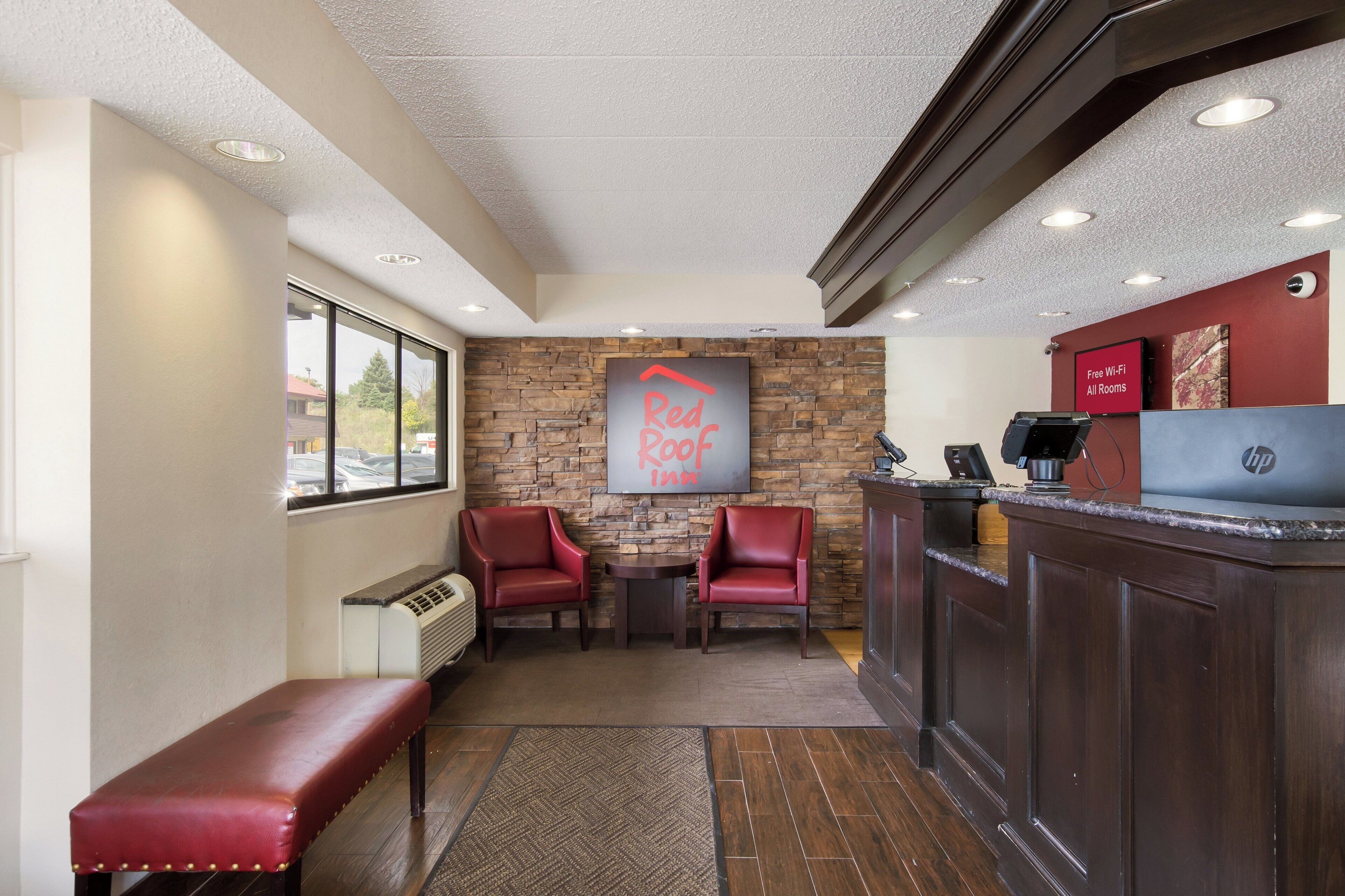 Una zona de recepción con pared de piedra, dos sillones rojos, un banco, un mostrador de recepción y un letrero que indica Wi-Fi gratuito en todas las habitaciones.