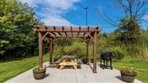 Eine hölzerne Pergola mit Picknicktisch und Grill in einem angelegten Garten.