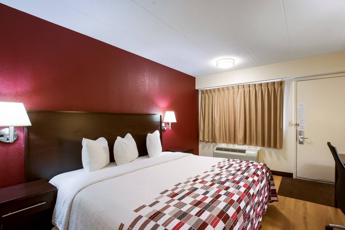 Une chambre d’hôtel avec un grand lit, une tête de lit en bois, une table de chevet, une lampe, une fenêtre avec des rideaux et une porte avec une pancarte.
