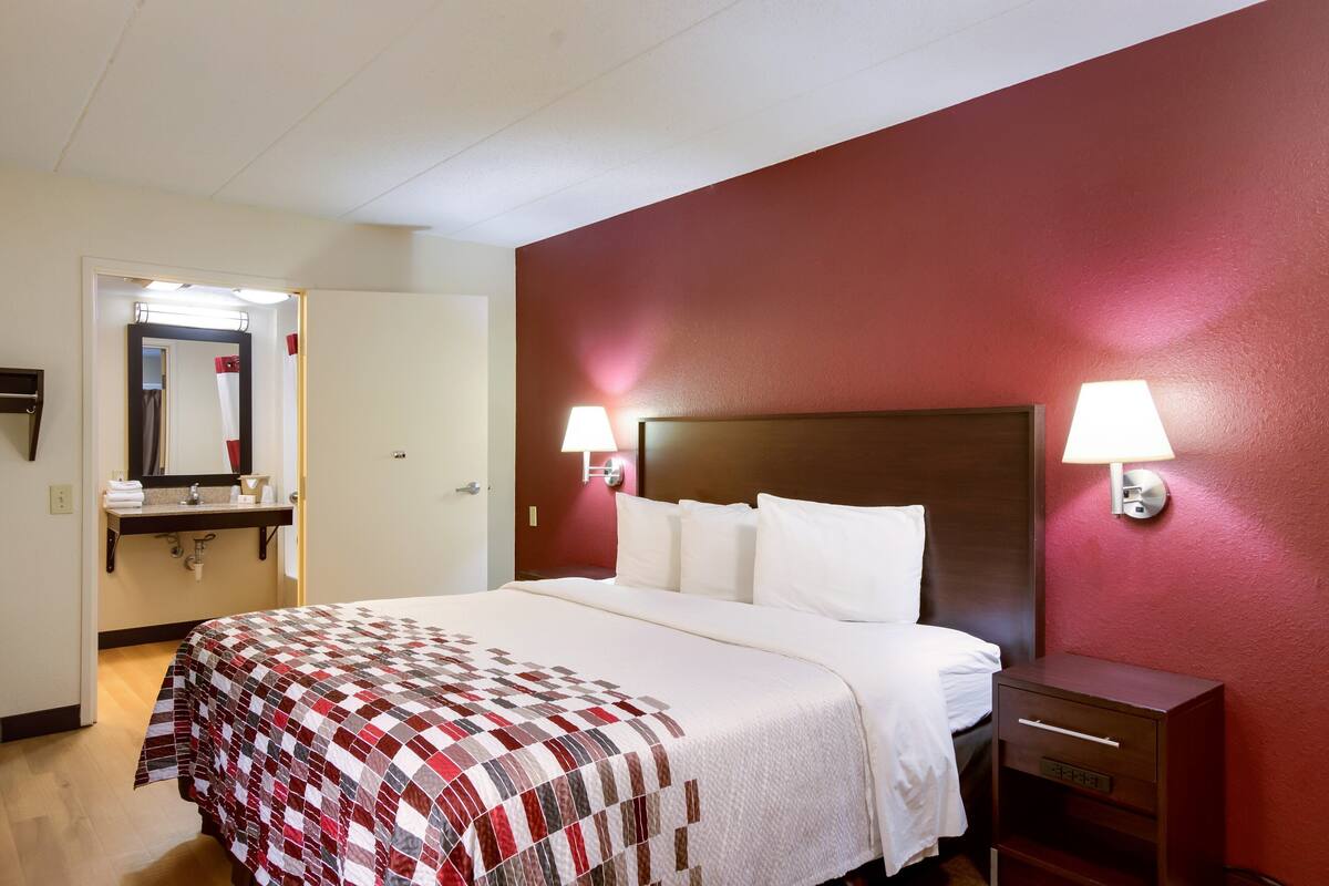 Une chambre d’hôtel avec un lit, une table de chevet, une lampe, un miroir et une salle de bain visible à travers une porte ouverte.
