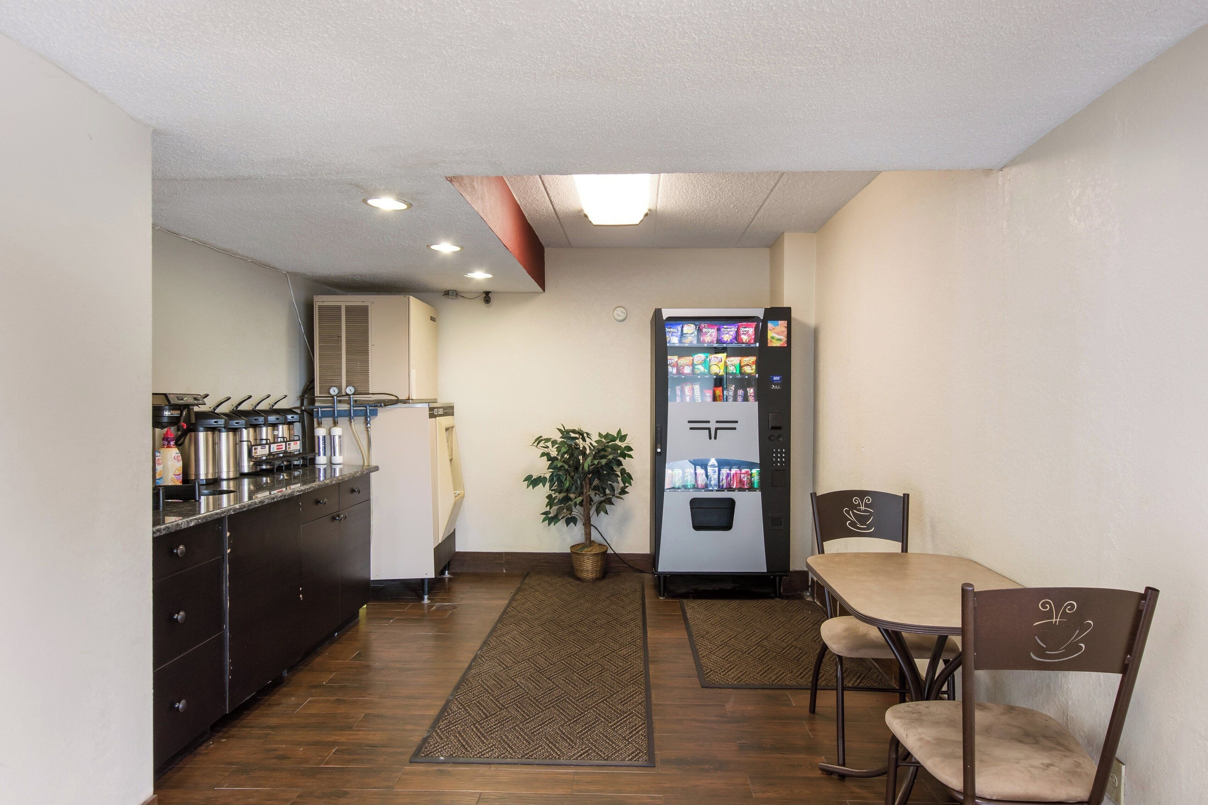 A compact hotel room with a kitchenette, a refrigerator, a dining area with a table and chairs, and a small potted plant.