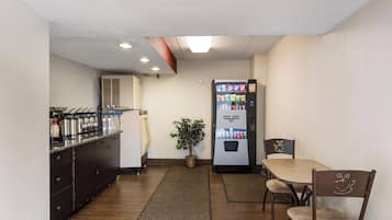A compact hotel room with a kitchenette, a refrigerator, a dining area with a table and chairs, and a small potted plant.