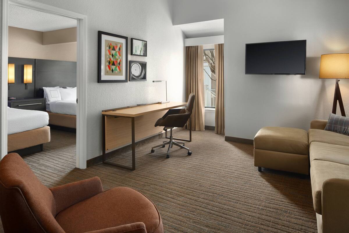 A hotel room with a bed, desk, chair, sofa, TV, and artwork on the wall.