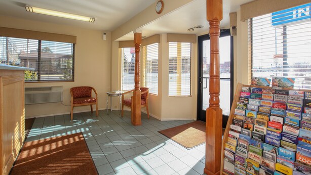 A hotel lobby with a reception desk, a wooden chair, a small table, a clock on the wall, and a bulletin board with travel brochures.