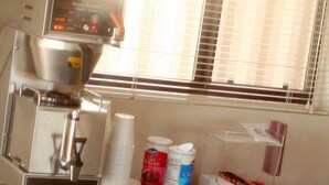 A coffee machine with a digital display, a window with blinds, and a counter with sugar, creamer, and a box of tissues.
