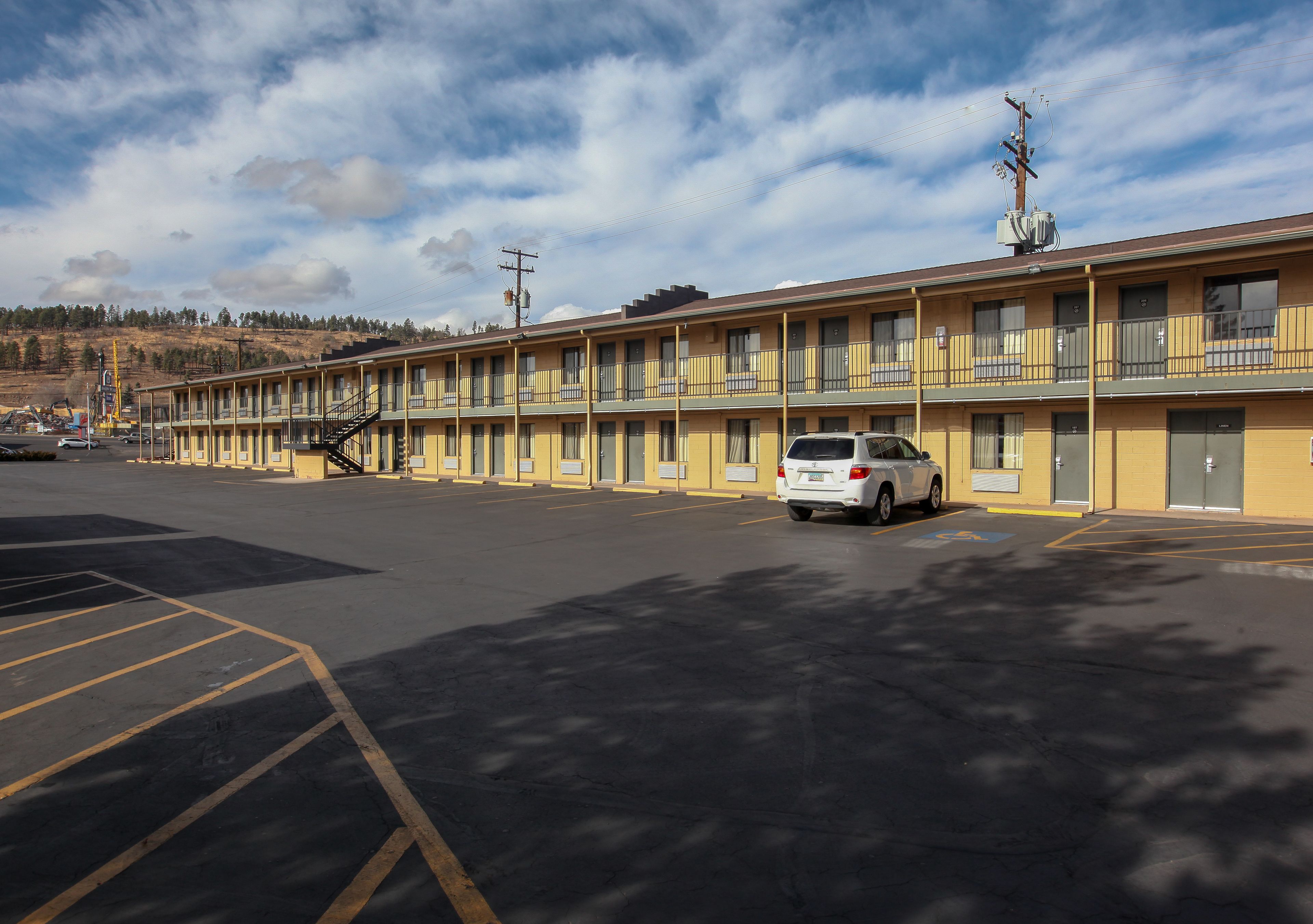 Un motel con una hilera de habitaciones, un estacionamiento y un paisaje montañoso al fondo.