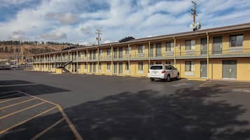 Un motel con una hilera de habitaciones, un estacionamiento y un paisaje montañoso al fondo.