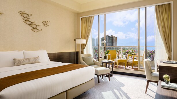 Een hotelkamer met een groot bed, een zithoek met een tafel en stoelen, en een balkon met uitzicht op de stad en gebouwen.