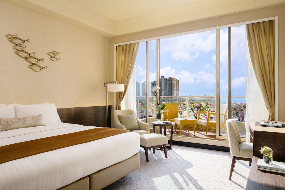 Une chambre d’hôtel dotée d’un grand lit, d’un coin salon avec une table et des chaises, et d’un balcon offrant une vue sur la ville et les bâtiments.