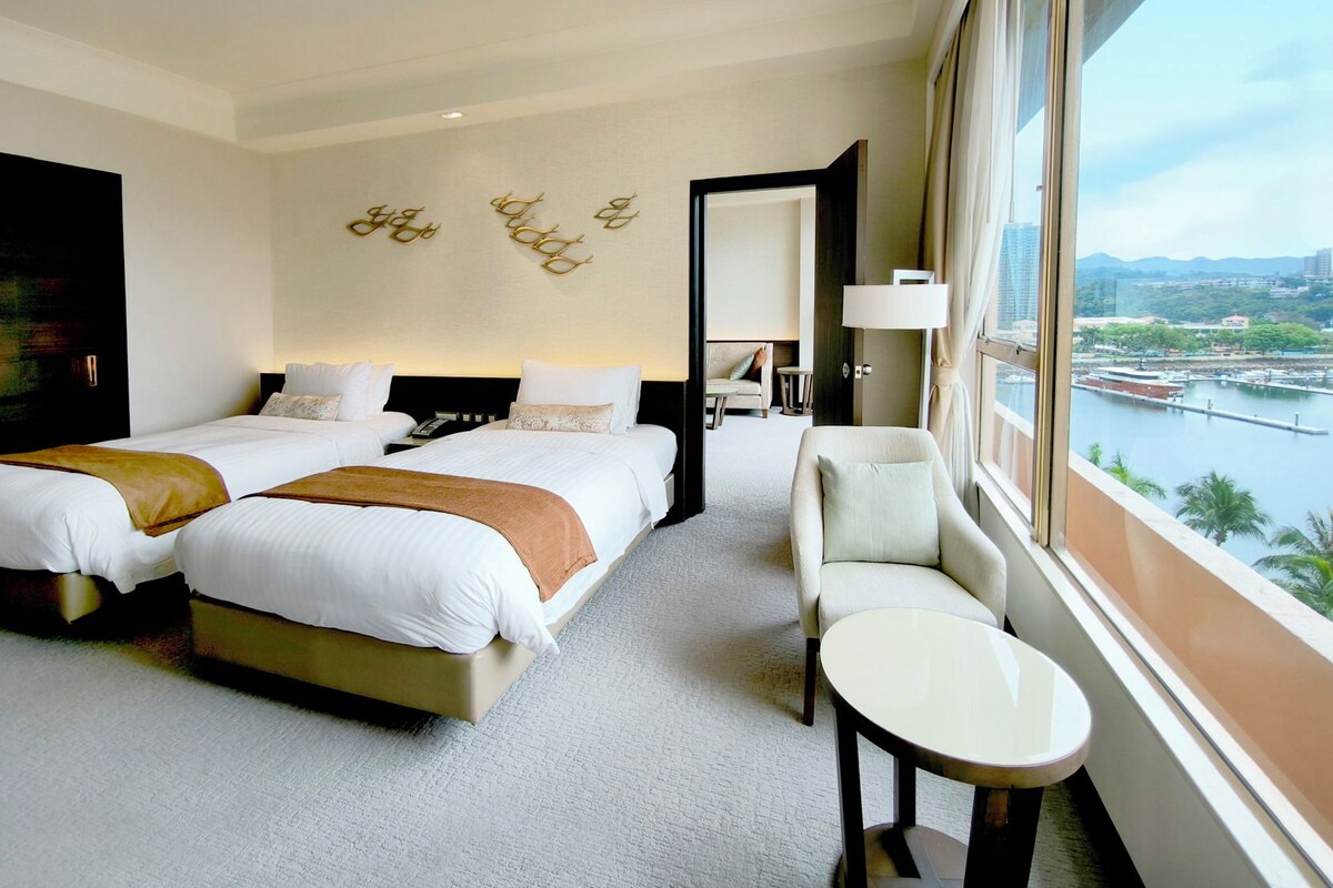 Een hotelkamer met twee bedden, een zithoek, uitzicht op een rivier en een stadsgezicht.