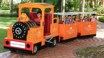 Un pequeño tren naranja con pasajeros, identificado como 'Gold Coast Hotel', circulando por un camino de ladrillo en un parque.
