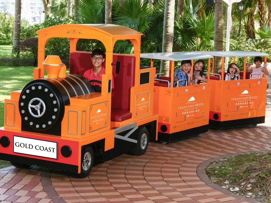Un pequeño tren naranja con pasajeros, identificado como 'Gold Coast Hotel', circulando por un camino de ladrillo en un parque.