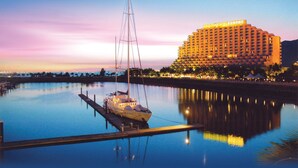 Een jachthaven met een boot afgemeerd aan een steiger, een groot hotelgebouw met verlichte reclame en rustig water dat de gebouwen weerspiegelt.