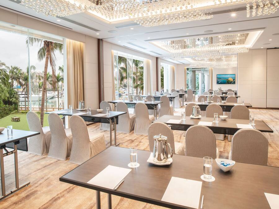 Een restaurantgedeelte in een hotel met verschillende tafels en stoelen, en een groot raam met uitzicht op palmbomen en een stadsbeeld.