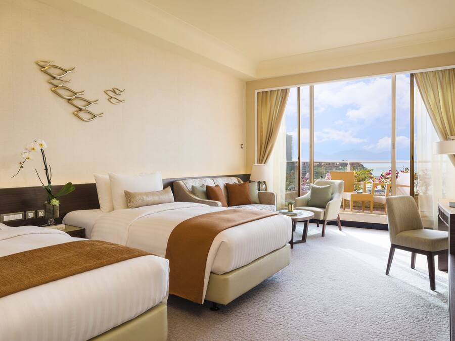 A hotel room with two beds, a desk, a chair, and a view of the city.