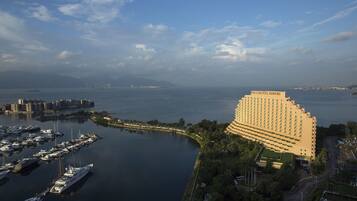 A large hotel with a curved facade, a marina with yachts, and a view of the sea and distant mountains.