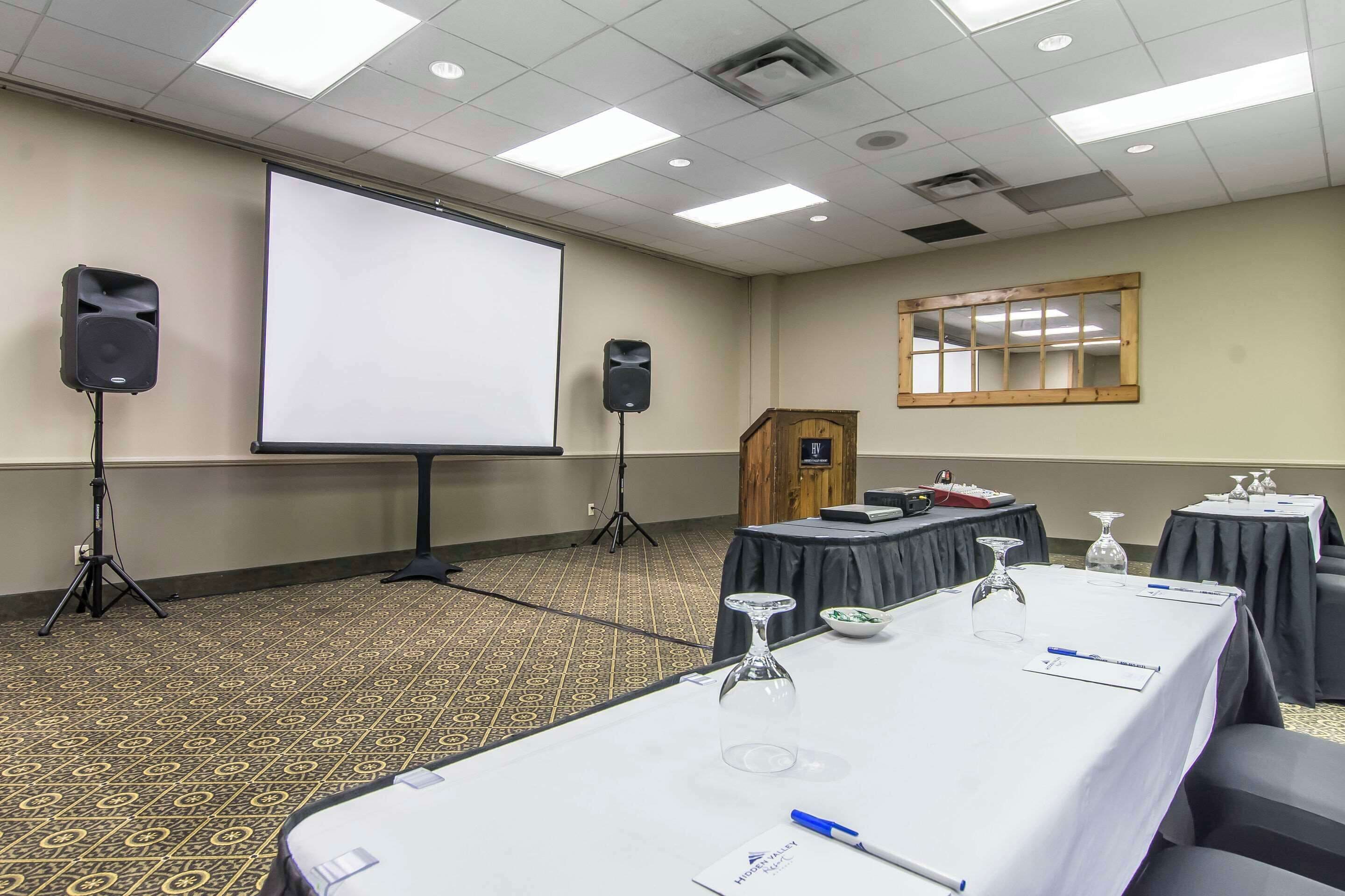 A conference room with a large projection screen, two speakers, a wooden podium, tables with nameplates, and a patterned carpet.