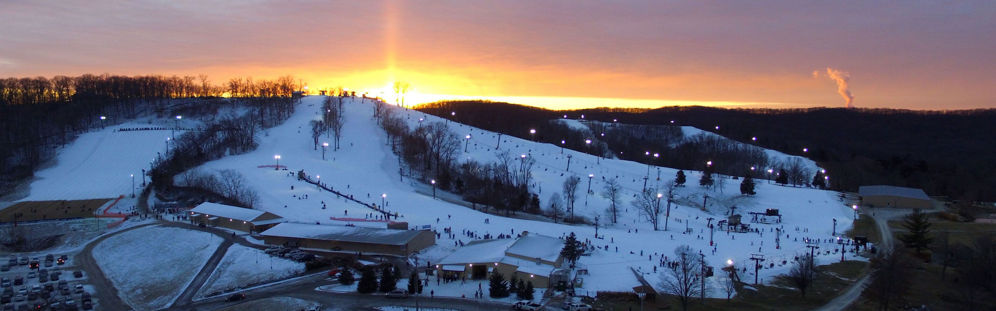 A ski resort at dusk with multiple ski lifts, a parking area, and buildings likely for lodging and dining.
