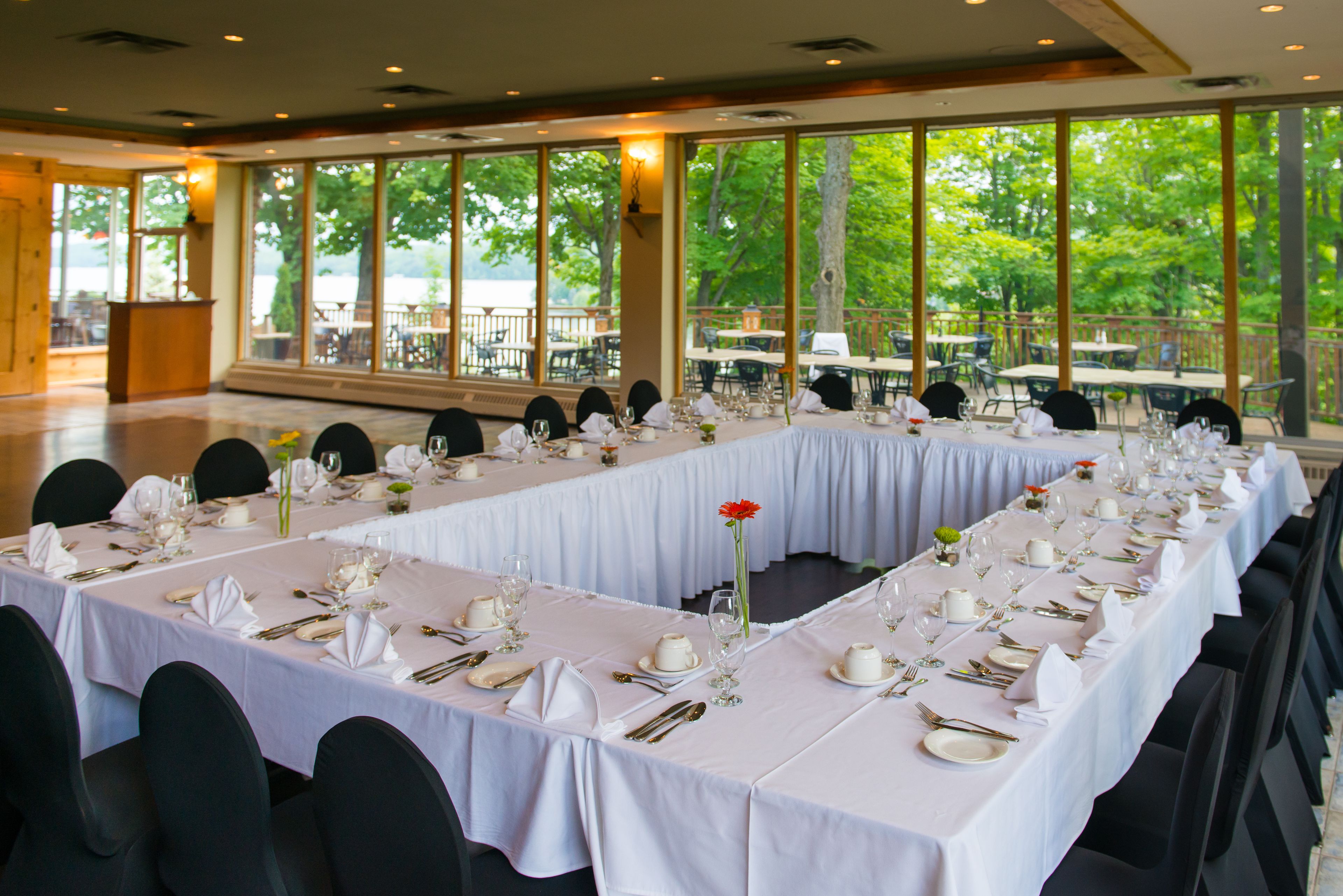 A banquet hall with long tables set for a formal event, large windows offering a view of the outdoors, and a podium at the front.