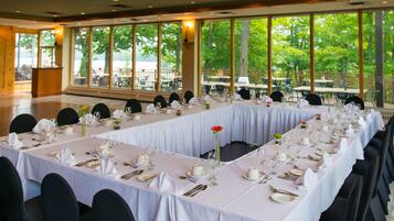 A banquet hall with long tables set for a formal event, large windows offering a view of the outdoors, and a podium at the front.