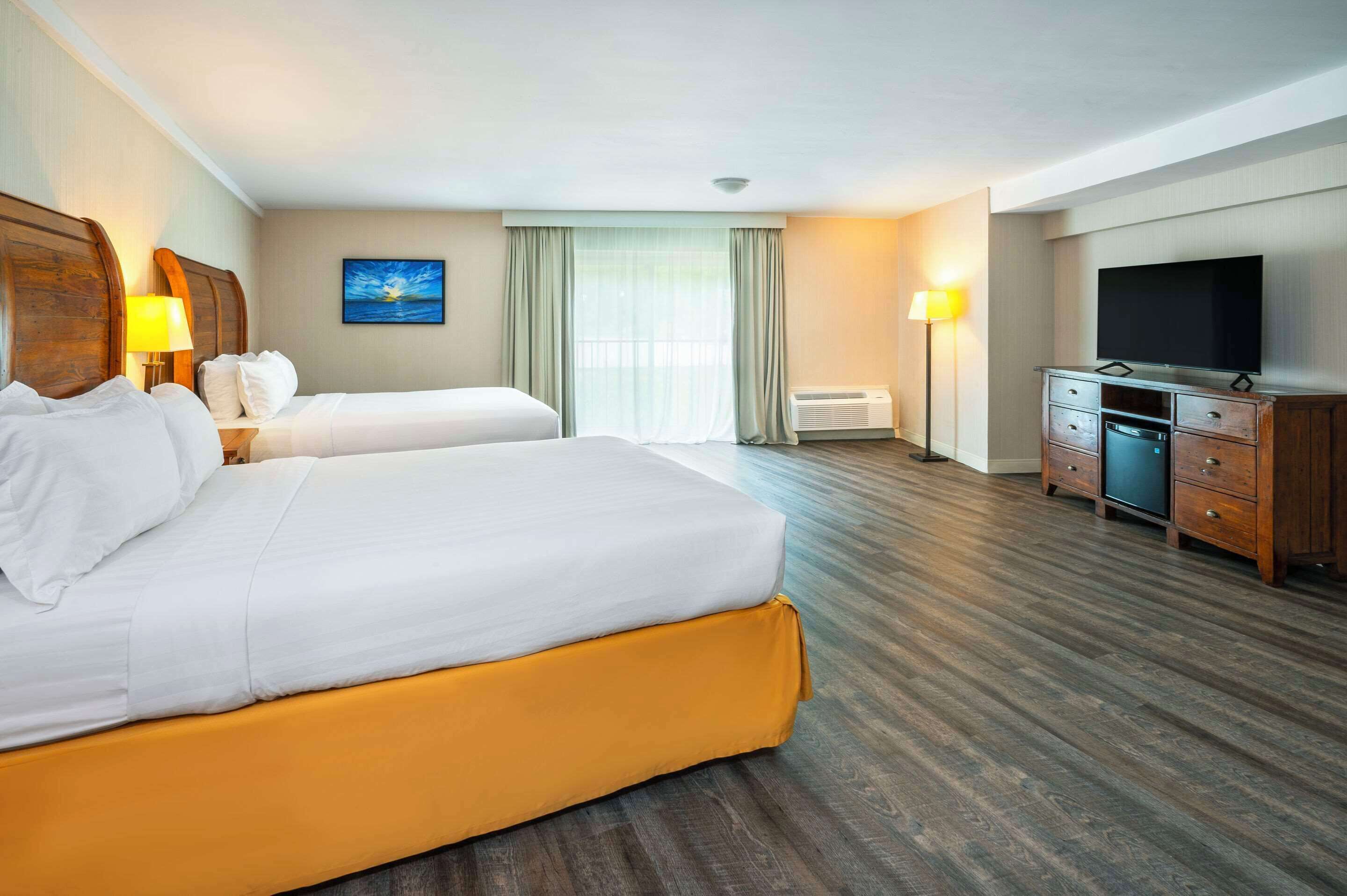 A hotel room with two beds, a flat-screen TV, a desk with a lamp, and a large window with curtains.