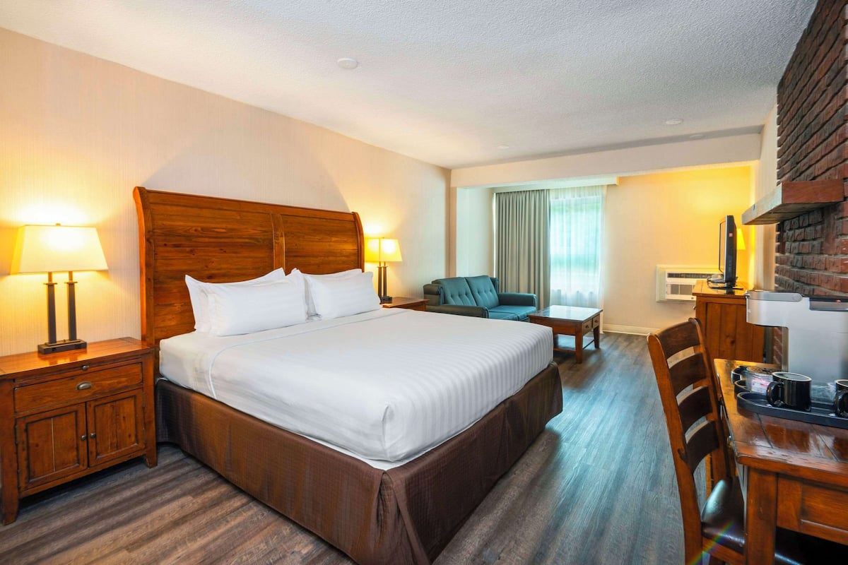 Habitación de hotel con una cama grande, dos mesitas de noche con lámparas, un sillón pequeño, una mesa de comedor con sillas y una chimenea de ladrillo.