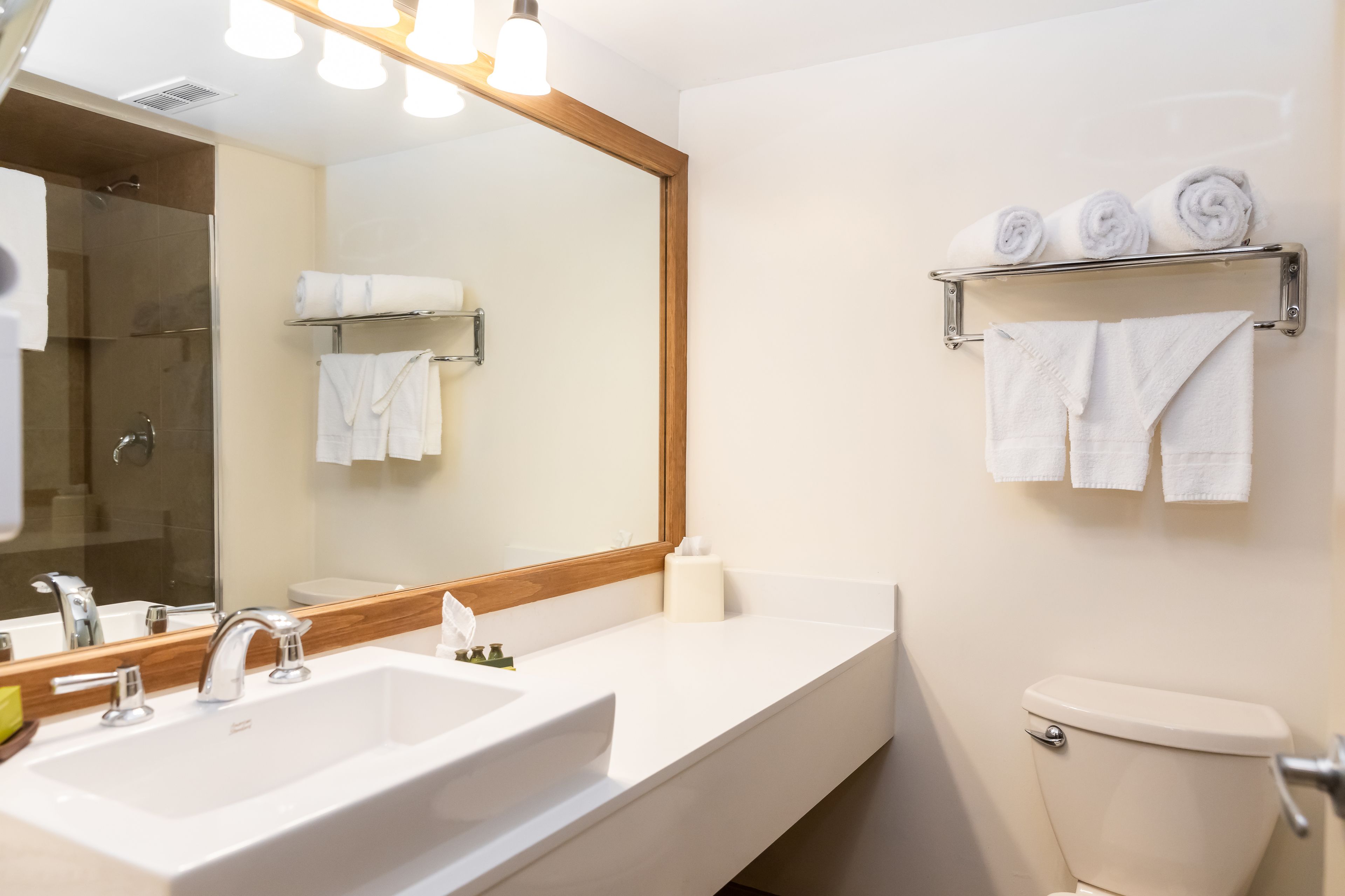 Un baño moderno con un espejo grande, un lavabo blanco y un inodoro. Hay toallas en un estante y una ducha se ve al fondo.