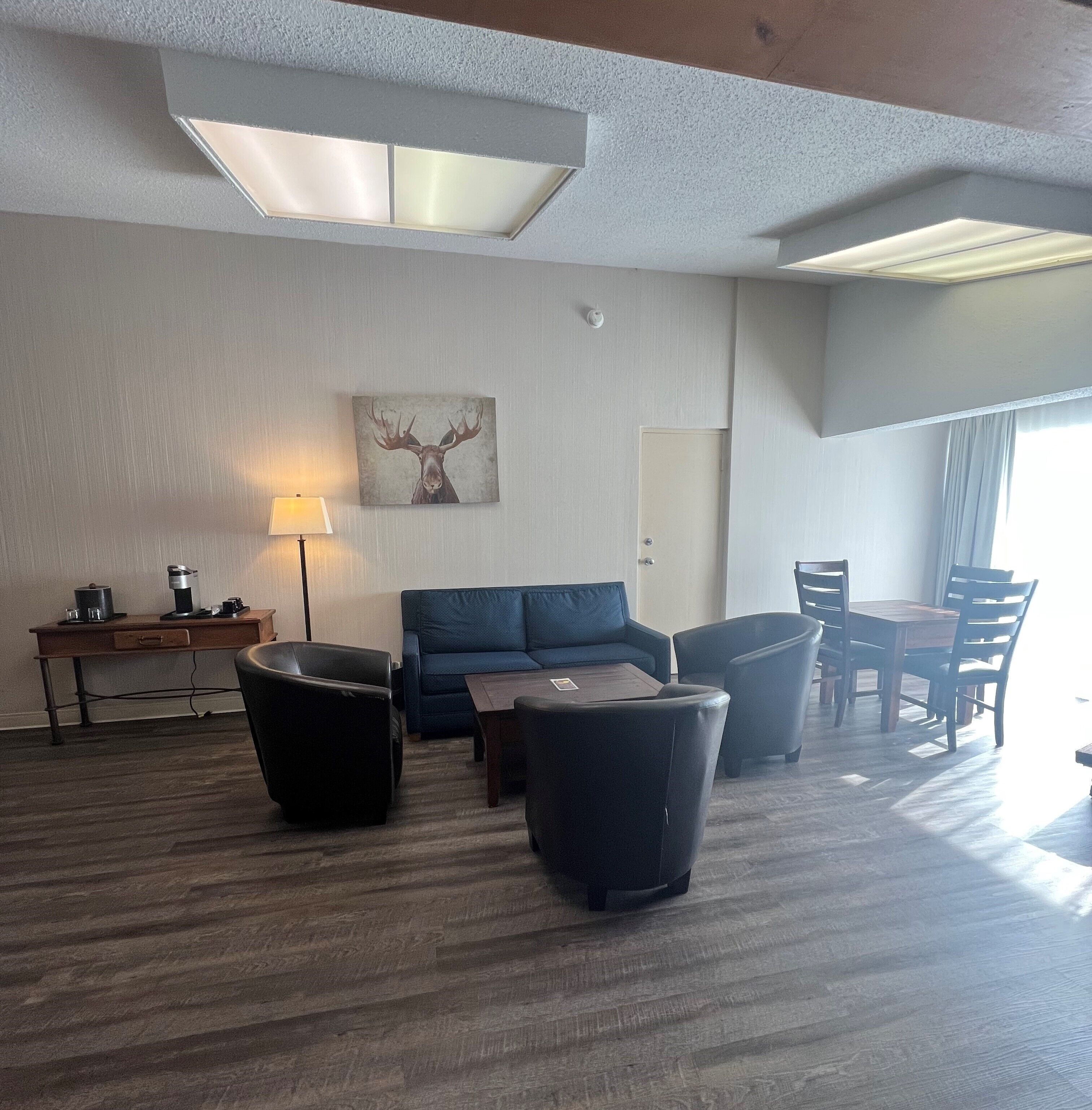 A modern living room with a sofa, two armchairs, a coffee table, a desk with a lamp, and a painting of a moose on the wall.