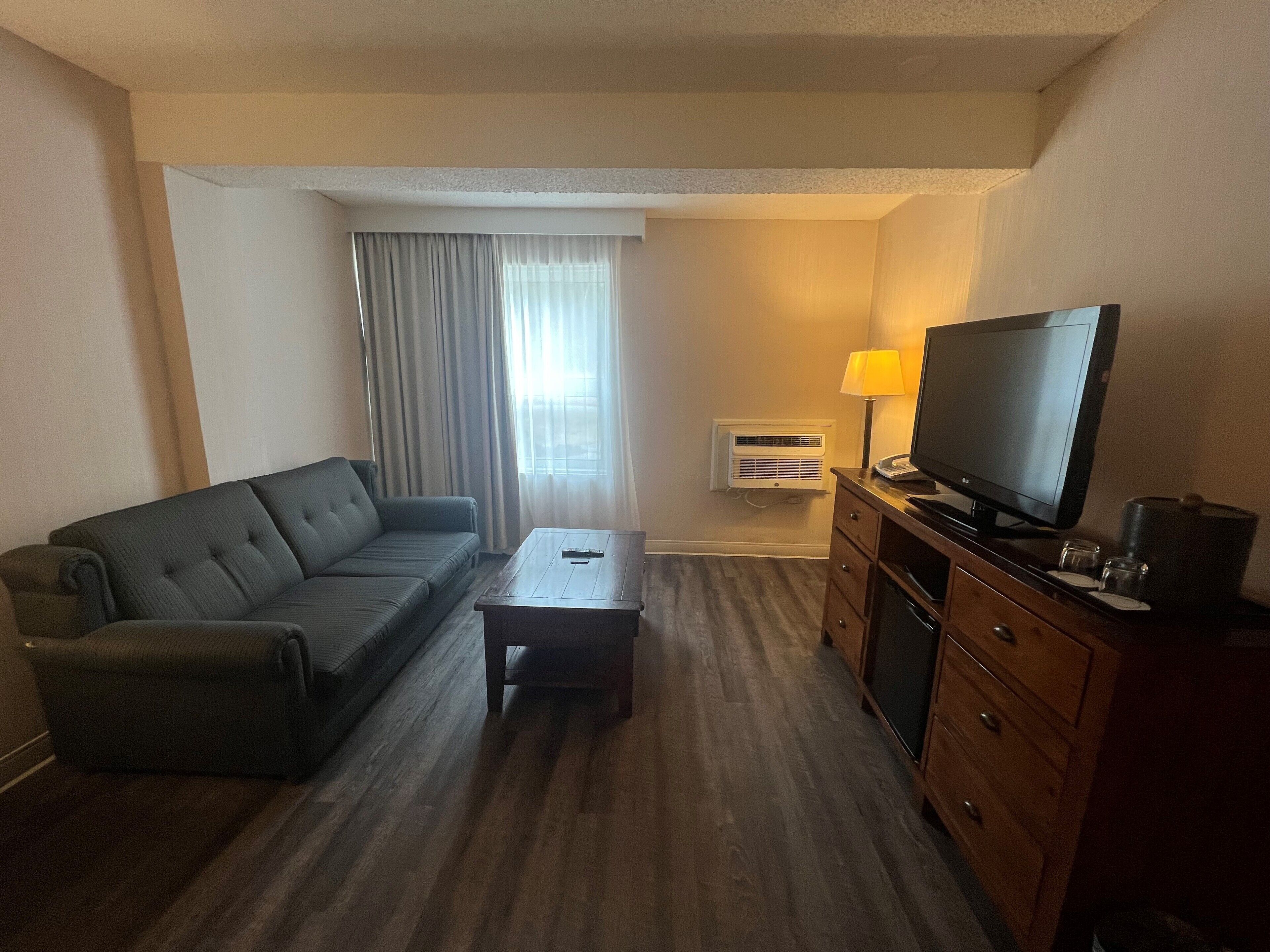 A hotel room with a dark green sofa, a wooden coffee table, a TV on a wooden cabinet, and a window with curtains.