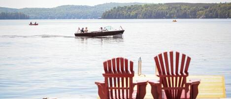A wooden dock with two Adirondack chairs facing a calm lake. Boats are visible on the water.