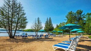 Ein Strand mit Reihen von Liegestühlen und Sonnenschirmen, ein Boot liegt am Wasser und Bäume im Hintergrund.