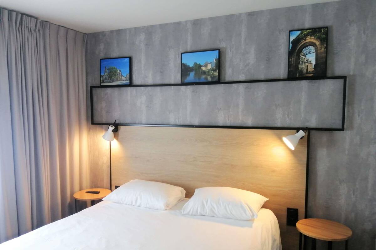 Une chambre à coucher avec un lit, deux lampes de chevet, une étagère murale ornée de tableaux et une table ronde.
