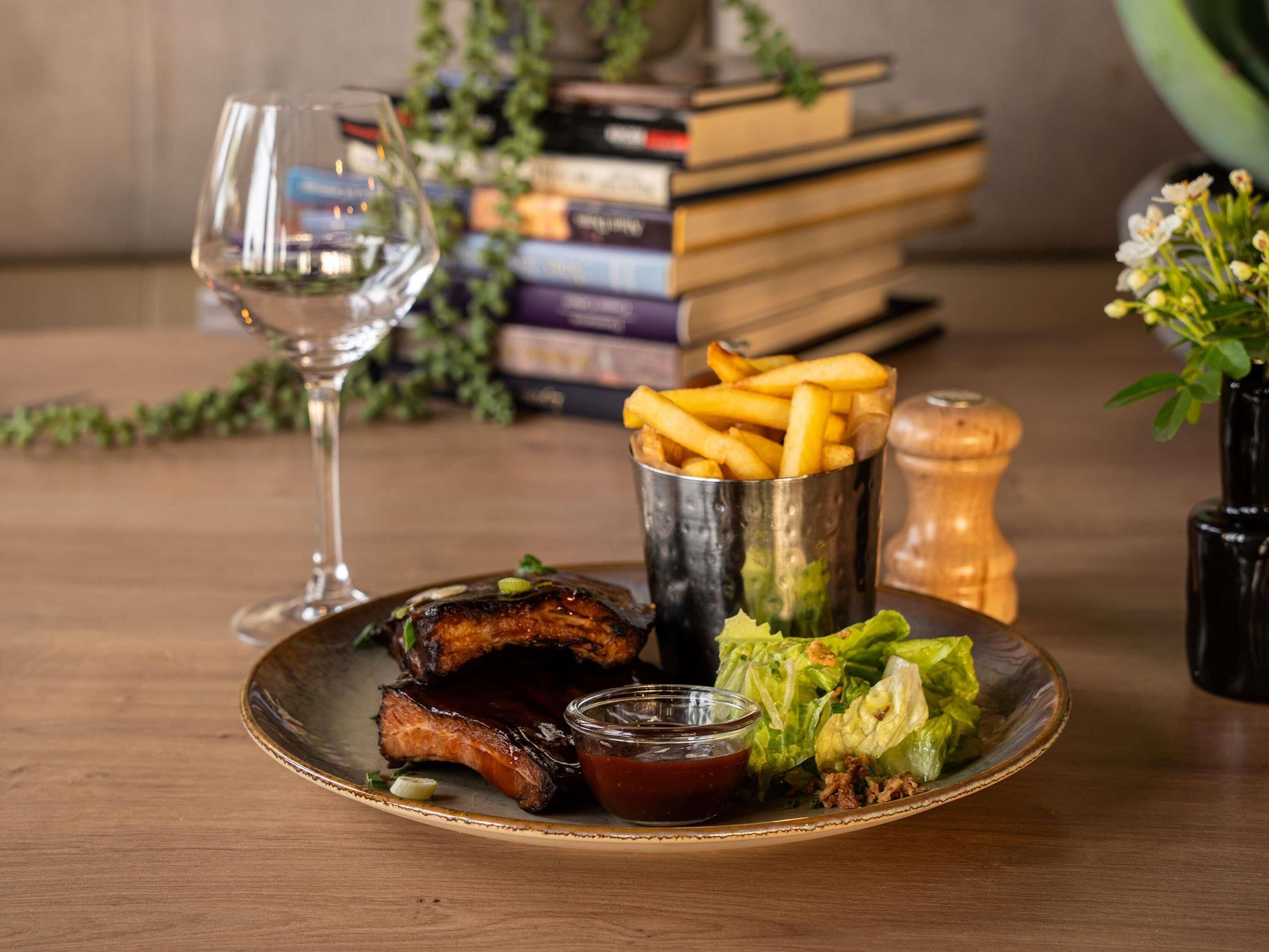 Een bord met ribben, friet en een salade, een glas wijn en een stapel boeken.