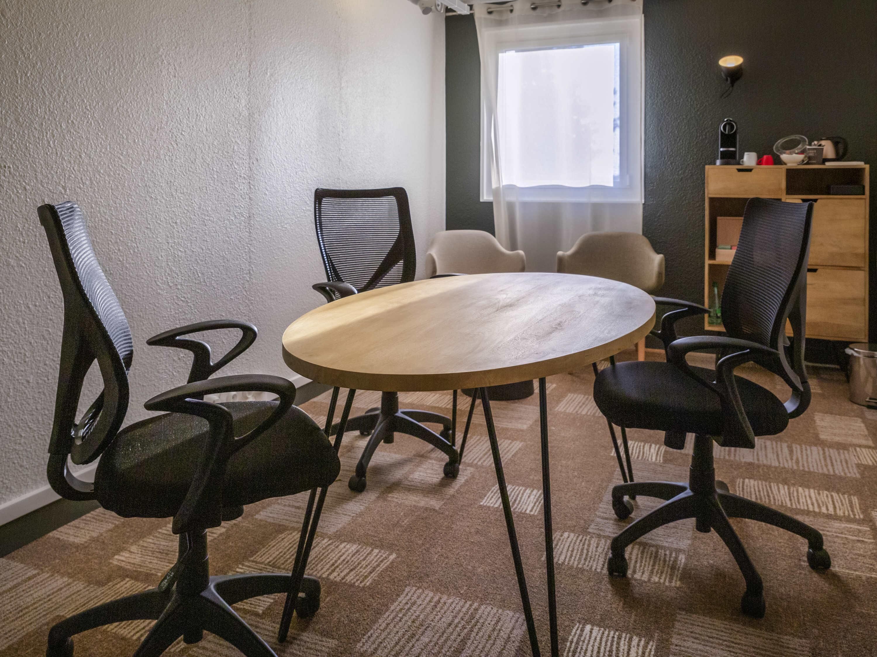 Une salle de conférence équipée d’une table ronde, de quatre chaises, d’un petit bureau avec un ordinateur et d’une fenêtre ornée de rideaux légers.