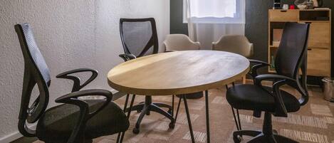 A conference room with a round table, four chairs, a small desk with a computer, and a window with sheer curtains.