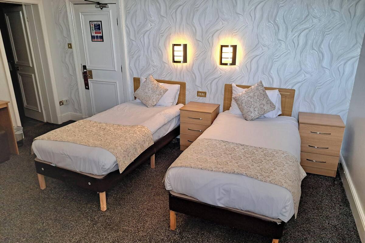 A hotel room with two single beds, wooden headboards, bedside tables with drawers, a white door, and a textured wall.