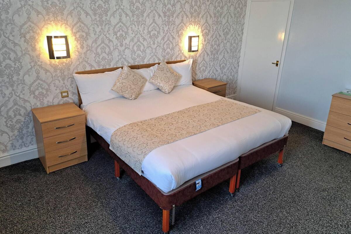 A neatly made bed with a wooden frame and a beige bedspread, flanked by two nightstands with drawers, in a room with patterned wallpaper and two wall-mounted light fixtures.