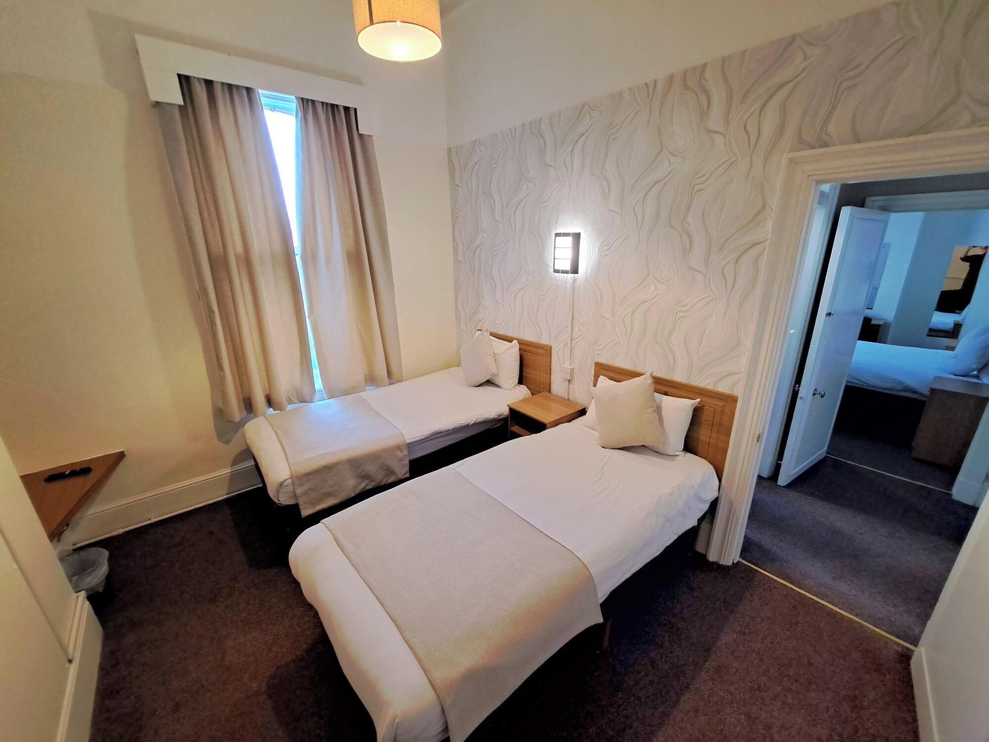 A hotel room with two single beds, a small bedside table, a nightstand, a lamp, and a patterned wall.