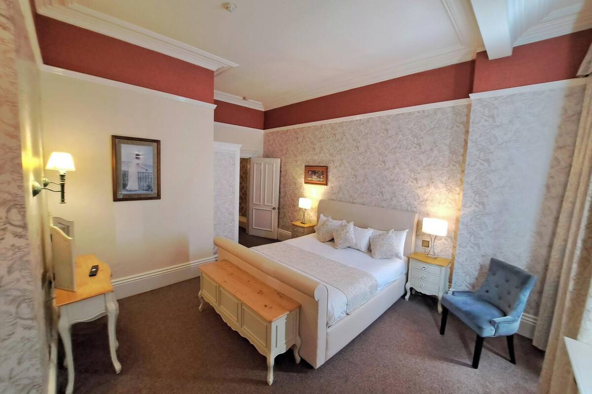 A hotel room with a large bed, two bedside tables with lamps, a wooden bench, a chair, and a wardrobe.