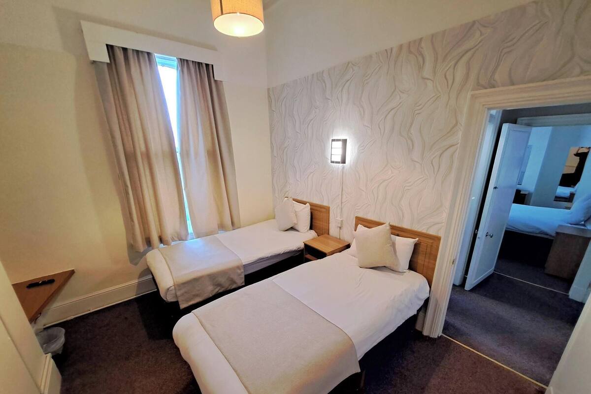 A hotel room with two single beds, a small bedside table with a lamp, a window with curtains, and a textured wall.