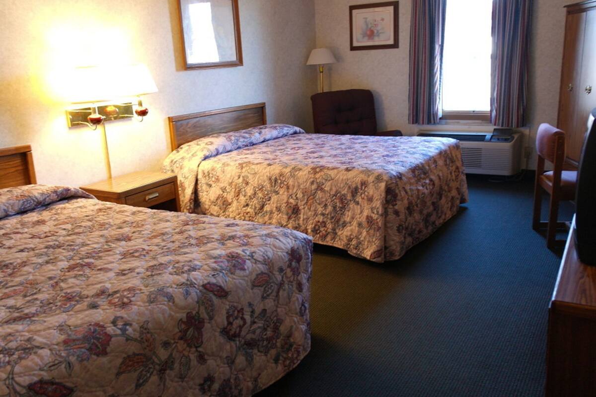 A hotel room with two beds, a desk, a chair, a lamp, an air conditioner, and a window with curtains.