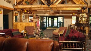 A cozy lodge interior with wooden beams, leather furniture, and a reception area with a wooden counter and glass doors. - Grand Ely Lodge Resort & Conference Center (Ely)