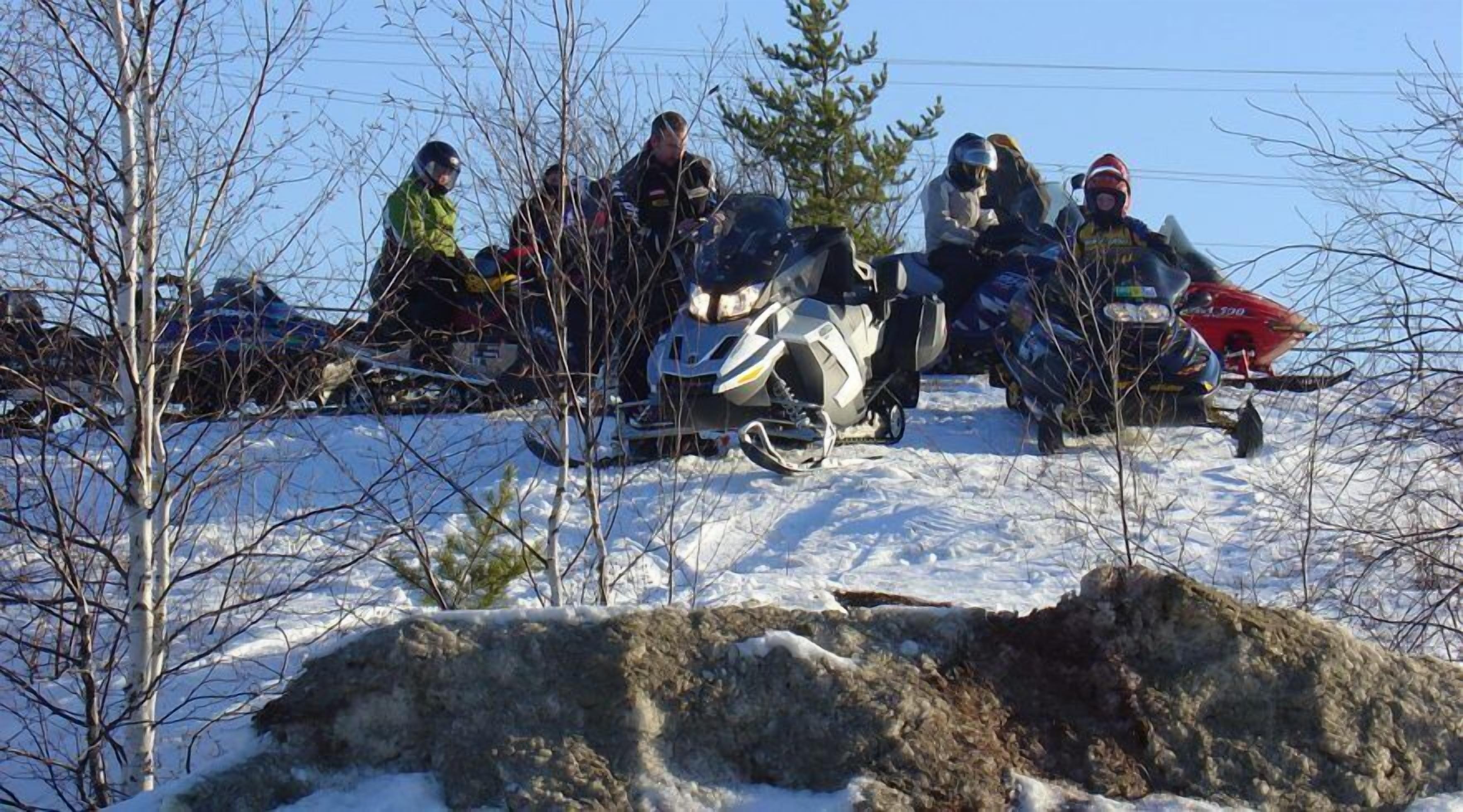 A group of people sitting on snowmobiles on a snowy slope with trees and rocks in the foreground.