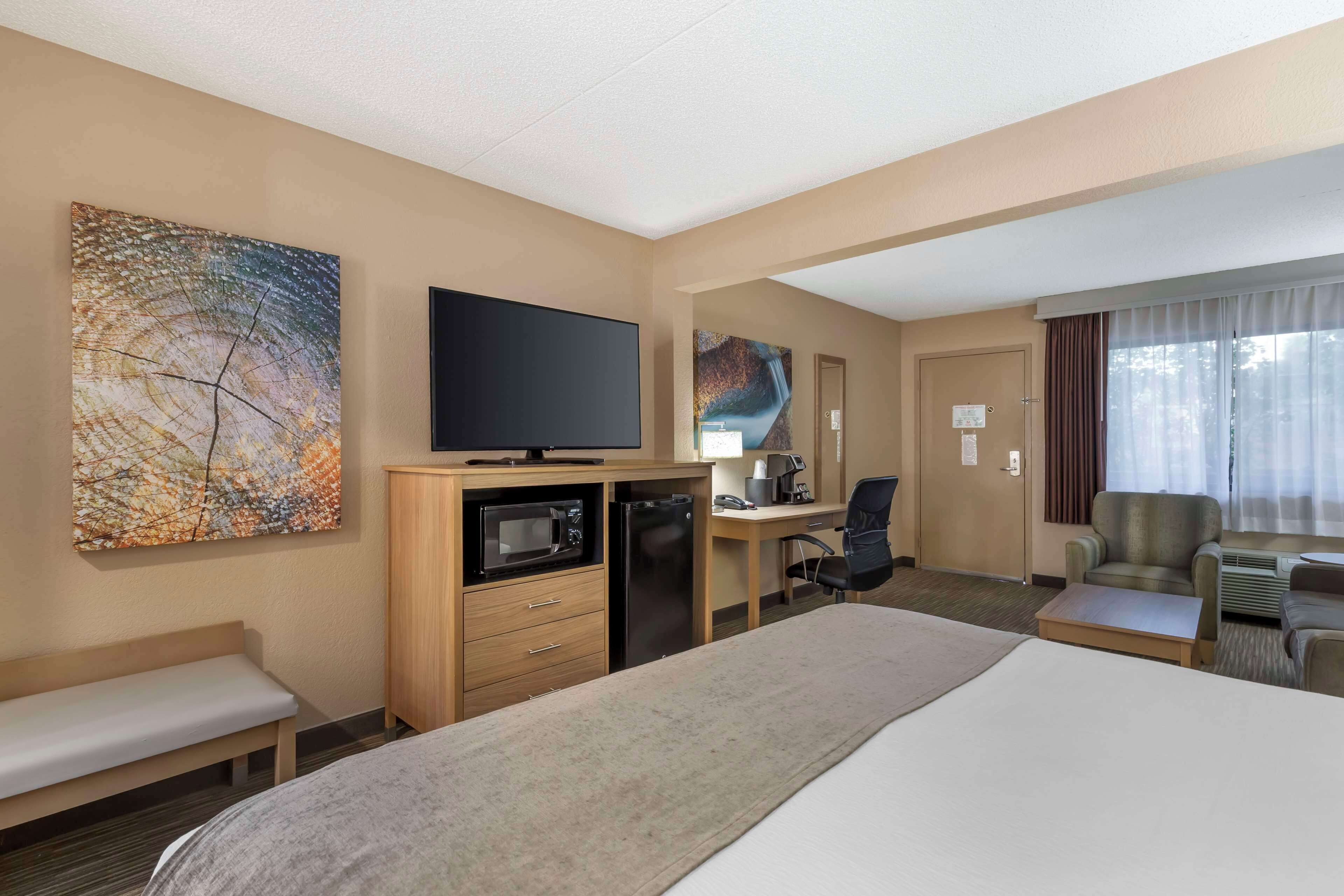 A hotel room with a bed, a desk, a chair, a TV, a microwave, a refrigerator, and artwork on the walls.