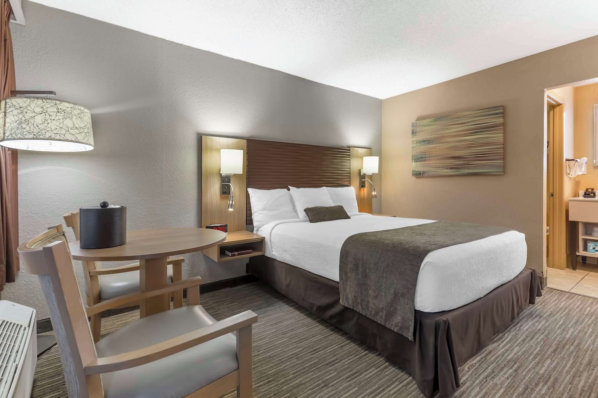 A hotel room with a bed, a desk, a chair, a lamp, a painting, and a bathroom visible through an open door.