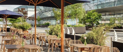 Zona de estar al aire libre con mobiliario de mimbre y sombrillas, con vista a un edificio moderno con ventanas de vidrio.