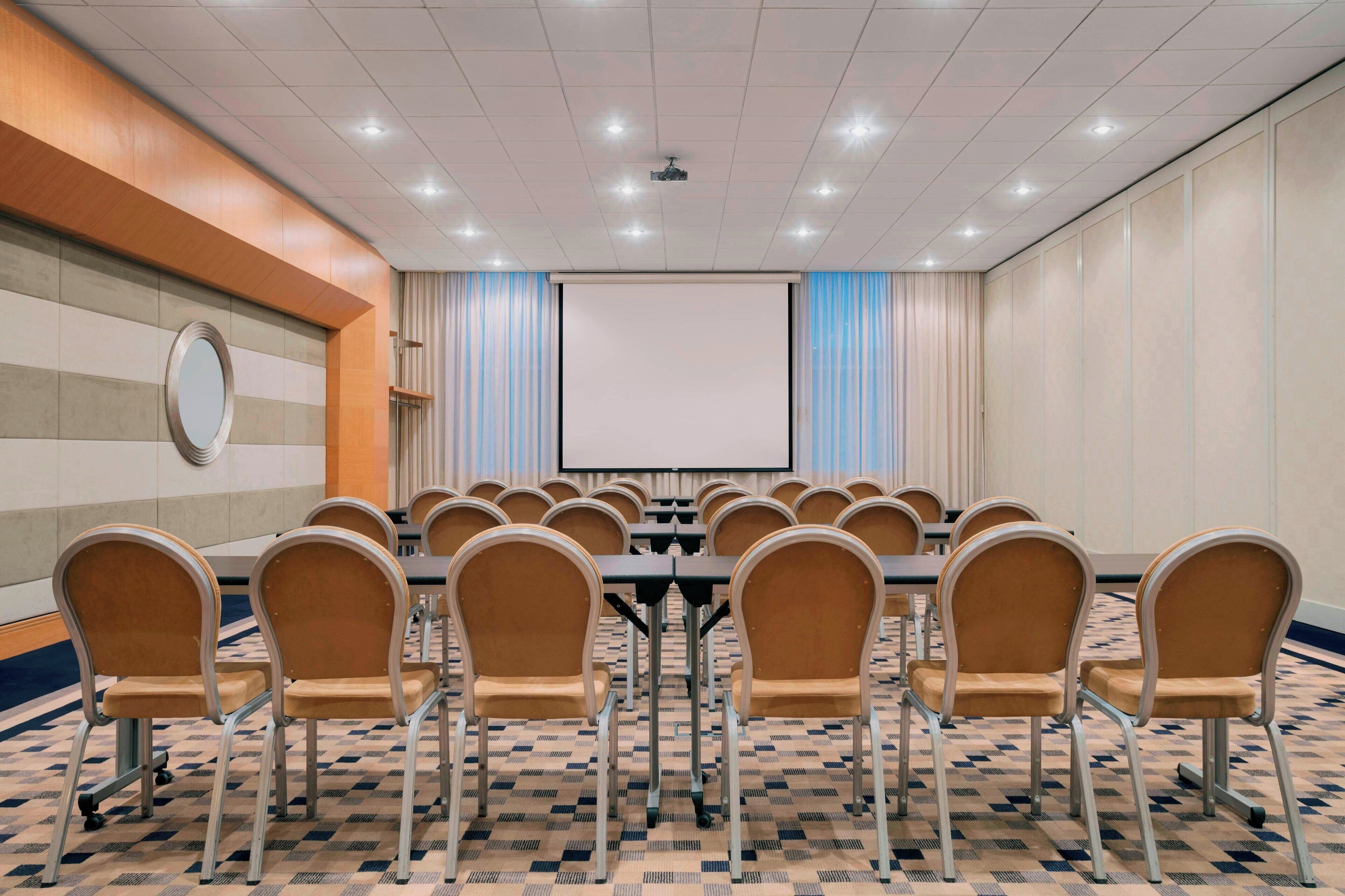 Una sala de conferències amb una taula gran, cadires, una pantalla de projector i una catifa estampada.