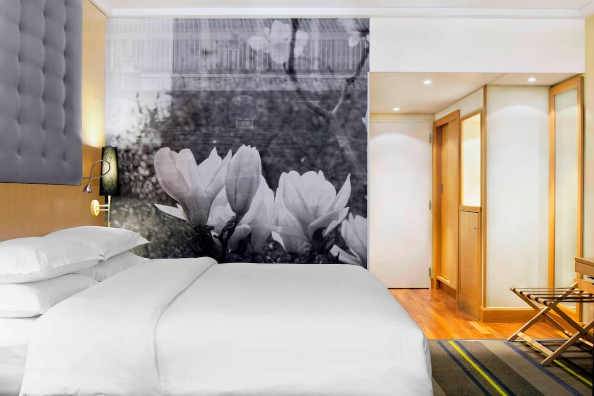 Une chambre d’hôtel avec un lit, une tapisserie murale représentant des fleurs, une tête de lit en bois, une table de chevet et une chaise pliante.