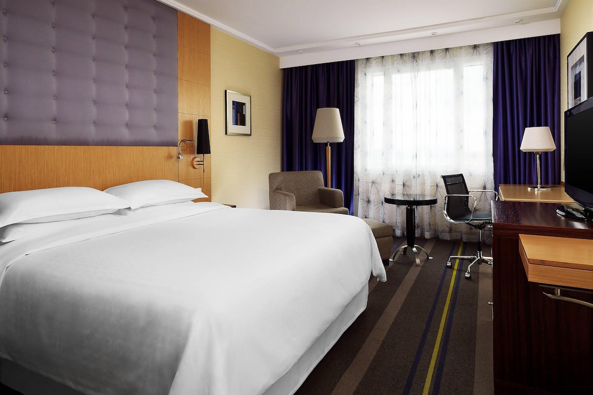 Une chambre d’hôtel avec un grand lit, un bureau avec une chaise, une télévision et une fenêtre avec des rideaux.