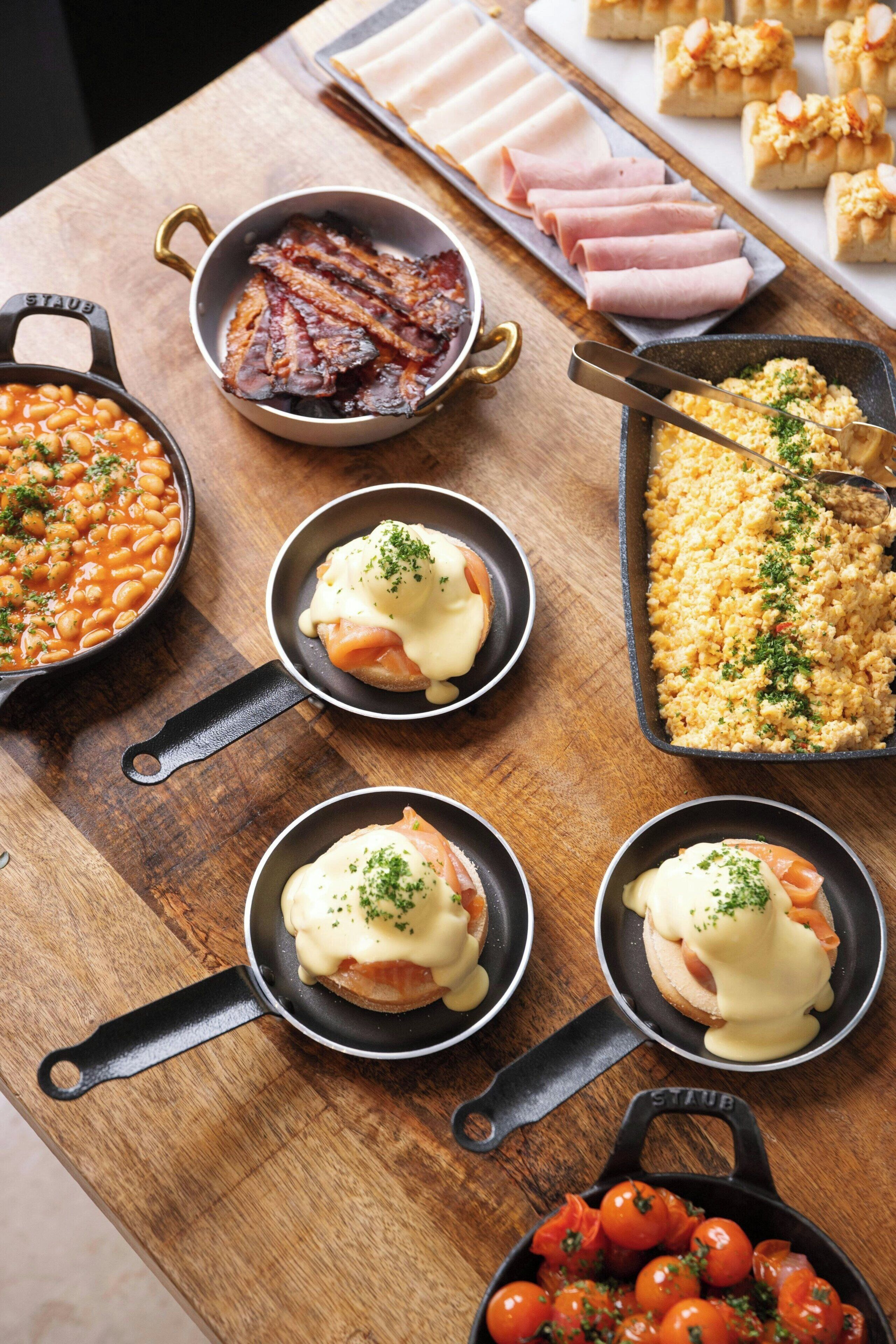 Une variété de plats sont disposés sur une table en bois, notamment des haricots cuits, du bacon et des œufs brouillés.